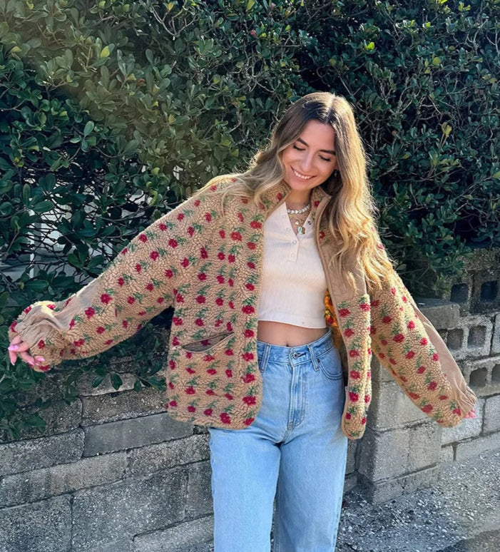 Woman wearing a floral cardigan and jeans standing against a stone wall with greenery.