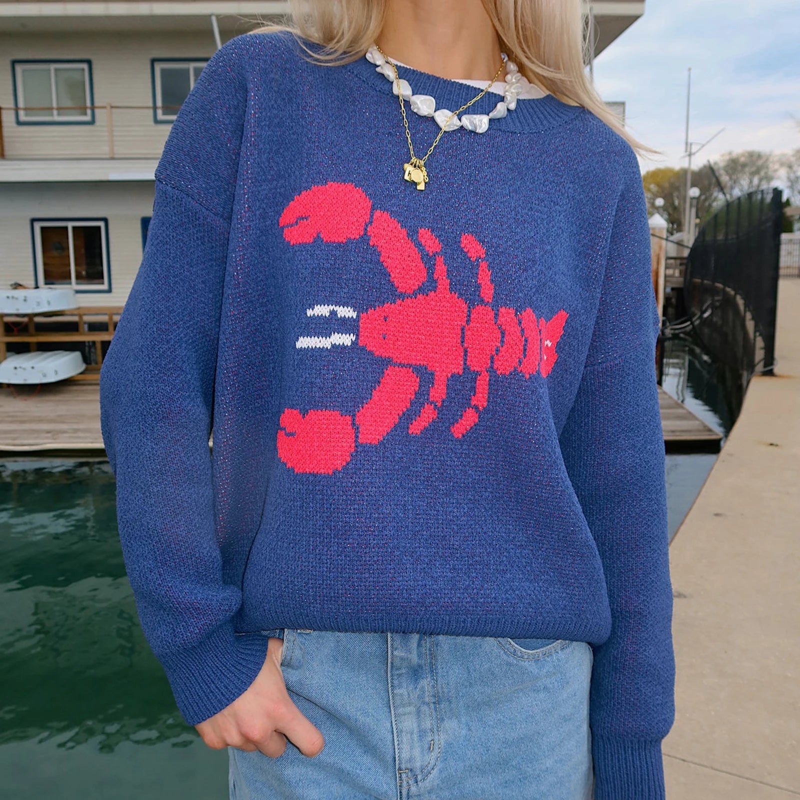 Person wearing a blue sweater with a red lobster design by a waterfront.