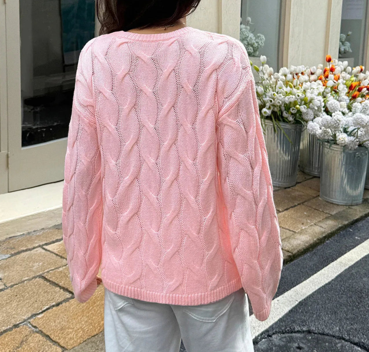 Person wearing a pink cable knit sweater standing on a sidewalk with flowers in the background.