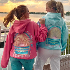 Two people wearing colorful hoodies with artistic designs and text, standing by a waterfront at sunset.