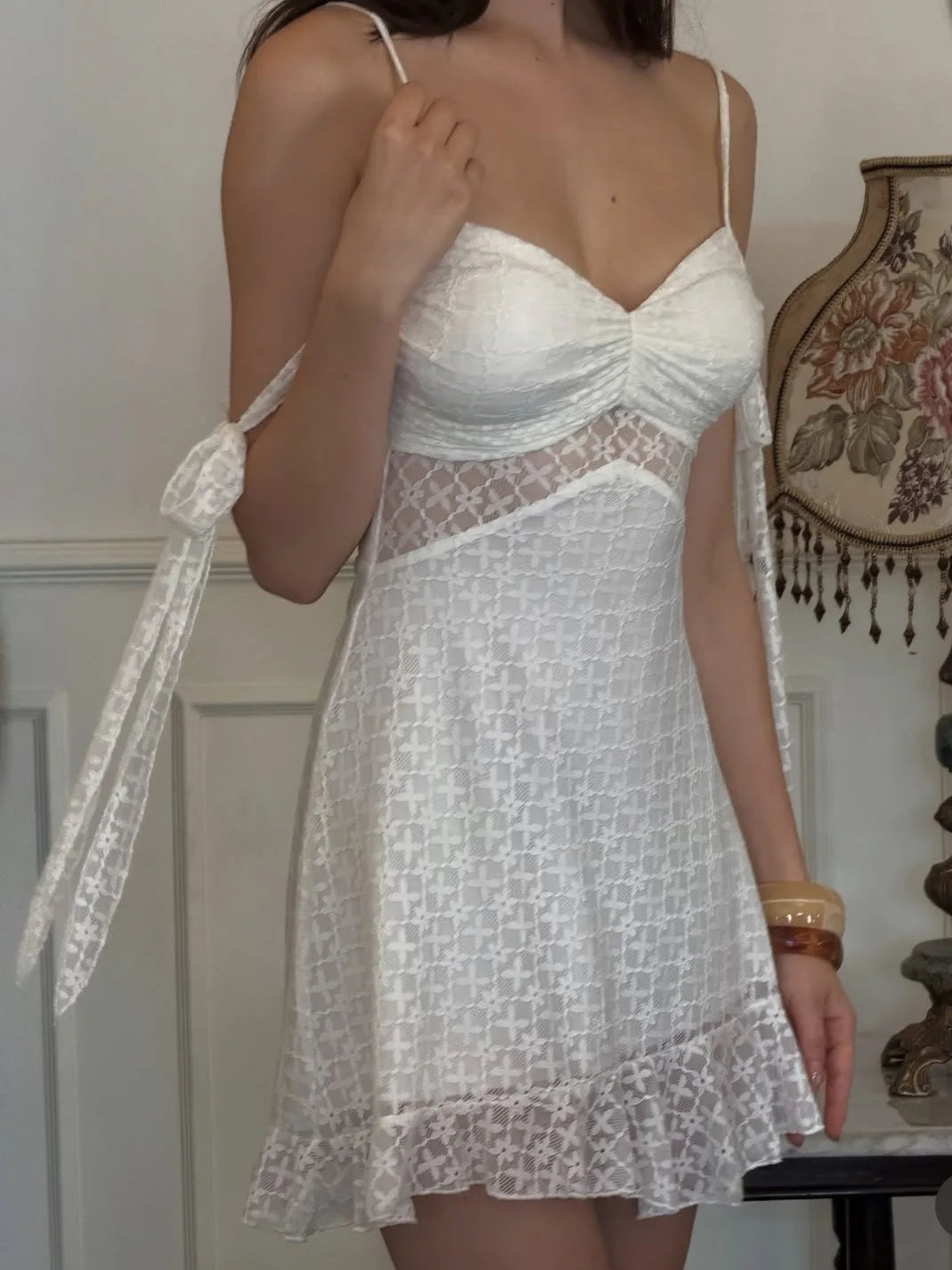 A woman wearing a white lace mini dress with thin straps, a ruched sweetheart neckline, sheer mesh panels, and a ruffled hem, standing indoors next to a decorative vase.