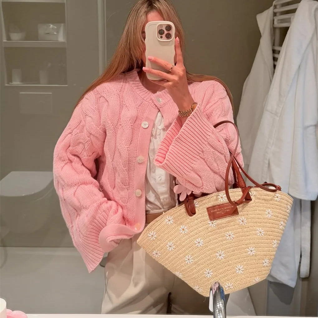 Person wearing a pink knitted cardigan holding a straw bag with floral patterns in a bathroom setting.