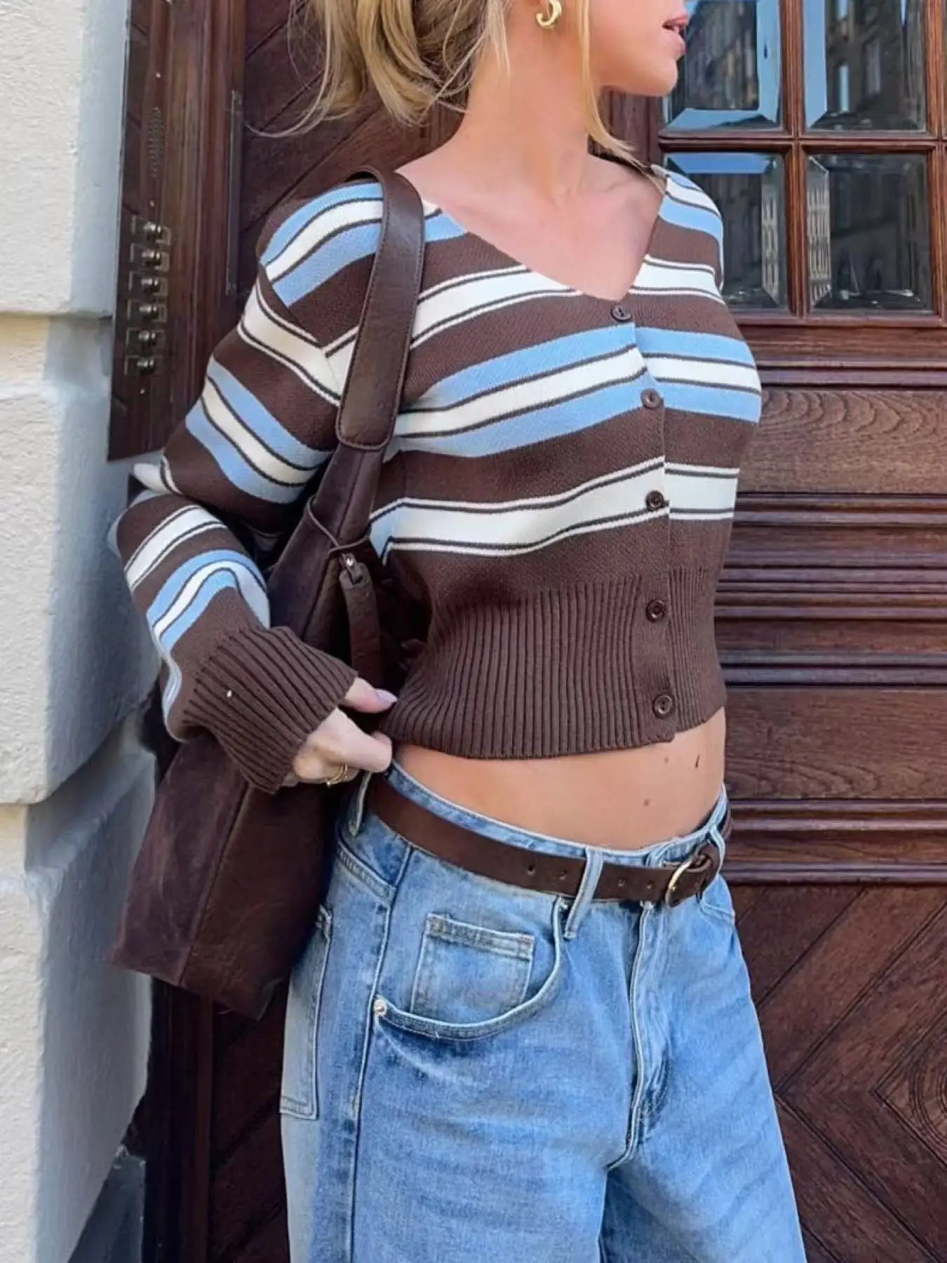 Person wearing a brown and blue striped sweater with high-waisted jeans, standing against a wooden door.