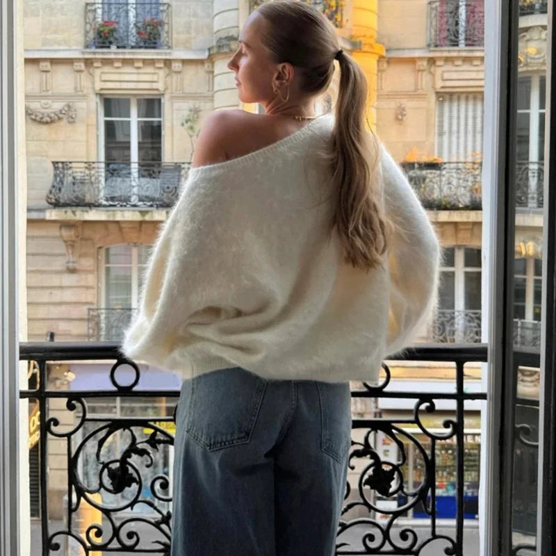 Woman in a white sweater and blue jeans standing on a balcony with a cityscape view.