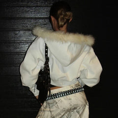 Person wearing a white hoodie with fur trim, holding a guitar, against a dark background.