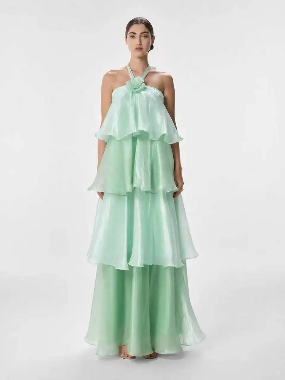 A woman modeling a mint green halter-neck maxi dress with a layered, tiered ruffle skirt and matching wide-leg pants against a plain light gray studio background.