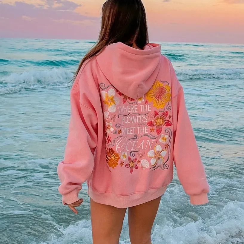 Person wearing a pink hoodie with floral design and text at the beach during sunset.