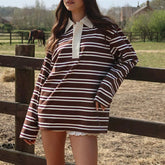 Person wearing a brown and white striped dress standing in a field with a horse in the background.