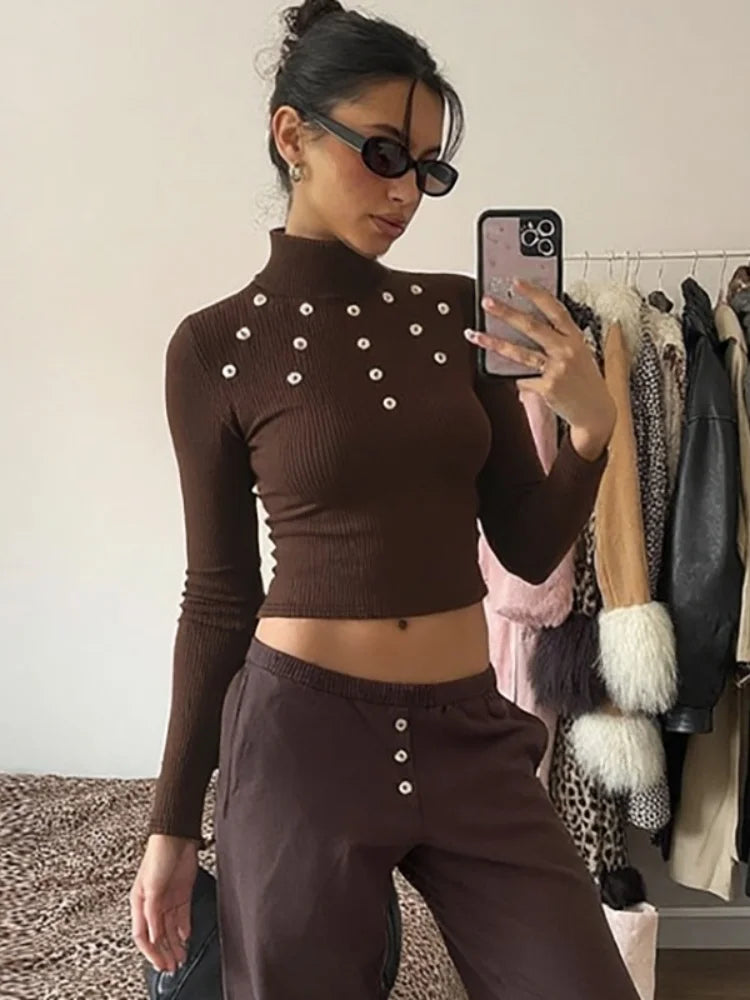 Person wearing a brown ribbed long-sleeve top and pants with button details, taking a mirror selfie in a room with clothing racks.