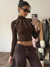 Person wearing a brown ribbed long-sleeve top and pants with button details, taking a mirror selfie in a room with clothing racks.