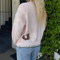 Person wearing a light pink sweater with a brown design, standing outdoors next to a building.