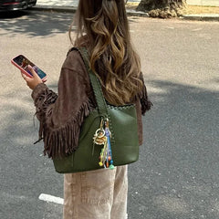 Person holding a phone and wearing a green bag with a fringed brown jacket outdoors.