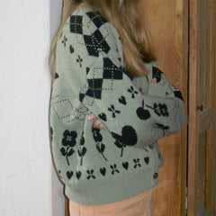 Person wearing a green sweater with black floral patterns, standing against a wooden door.