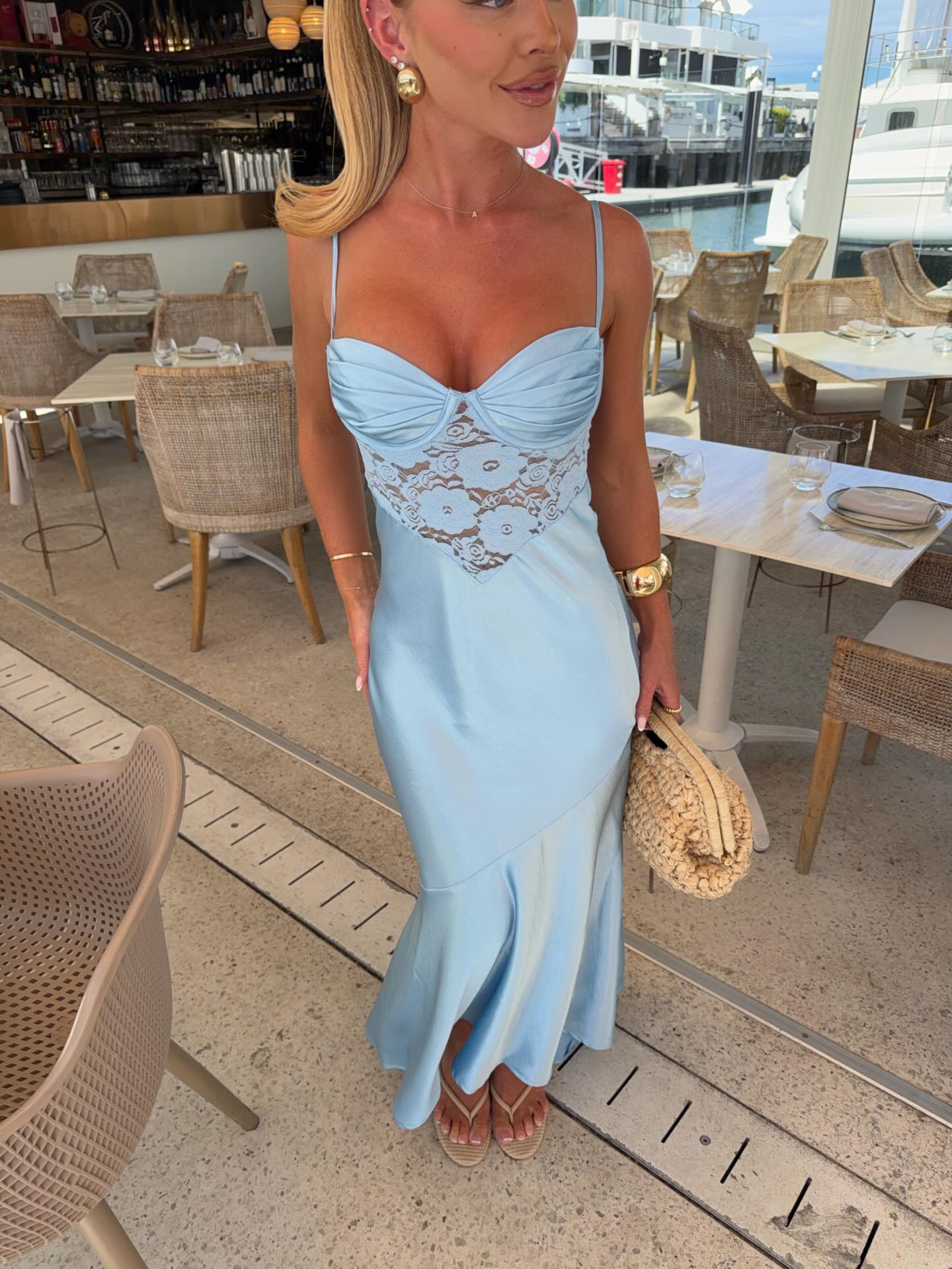 A woman wearing a light blue satin maxi dress with a lace corset-style bodice, standing in a marina restaurant setting.