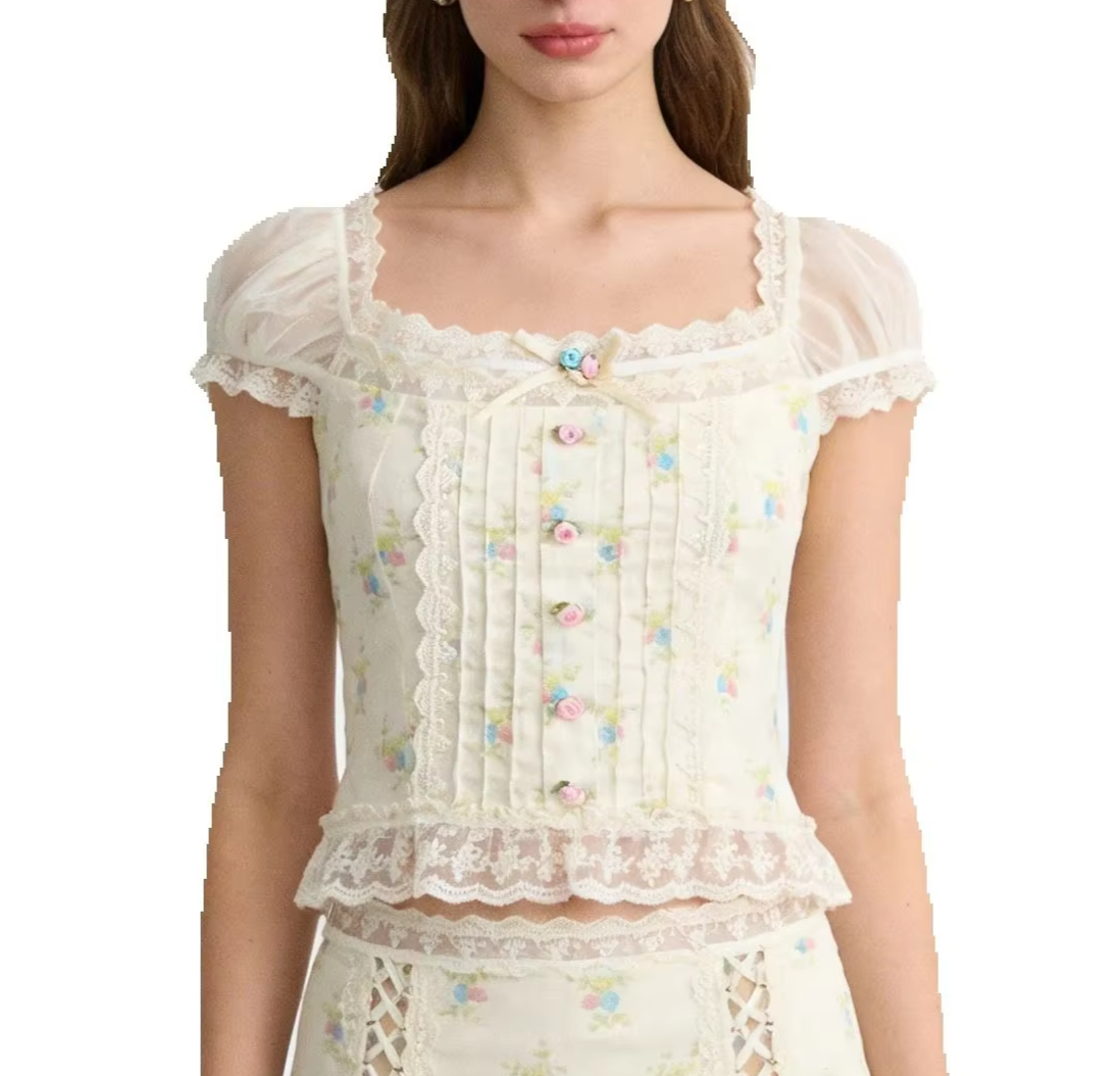 A model wearing a white lace-trimmed blouse with a delicate pastel floral print, featuring a square neckline, sheer puff sleeves, and decorative buttons down the front.