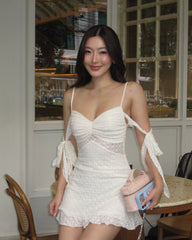 A woman wearing a white lace mini dress with thin straps, a ruched sweetheart bodice, and tie details on the sleeves, holding a pink and blue clutch bag in an indoor cafe setting.