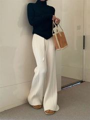 Person wearing a black turtleneck and white wide-leg pants, holding a brown bag indoors.