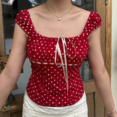 Person wearing a red polka dot top with a bow detail, standing indoors.