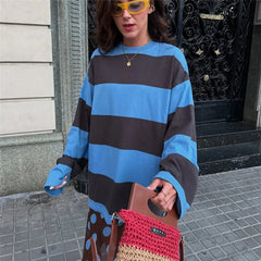 Person wearing a blue and brown striped sweater holding a red and brown bag on a sidewalk.
