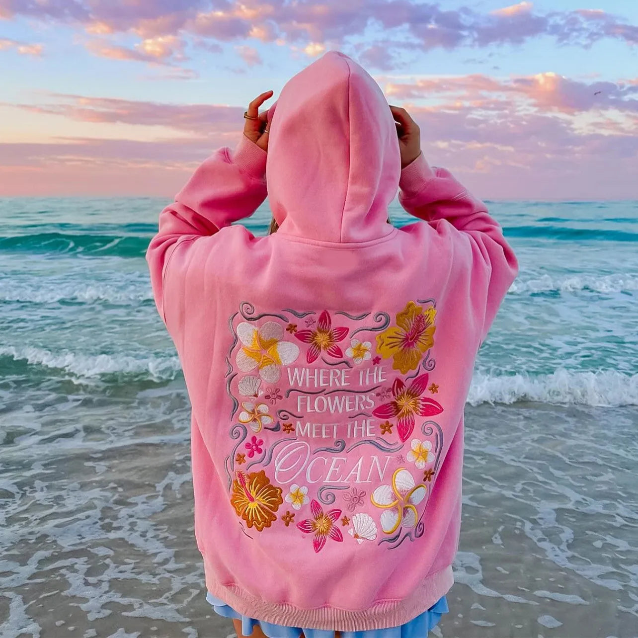 Person wearing a pink hoodie with floral design and text at the beach during sunset.