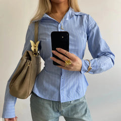 Person wearing a blue striped shirt holding a phone, with a beige handbag on a plain background