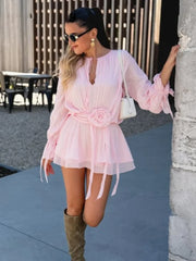 Woman wearing a pink dress with a white bag and sunglasses, standing against a wall.