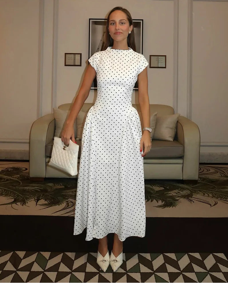 Woman in a white polka dot dress standing in an elegant room with a sofa and framed pictures.