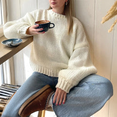 Person wearing a cream sweater and blue jeans, holding a mug in a cozy indoor setting.