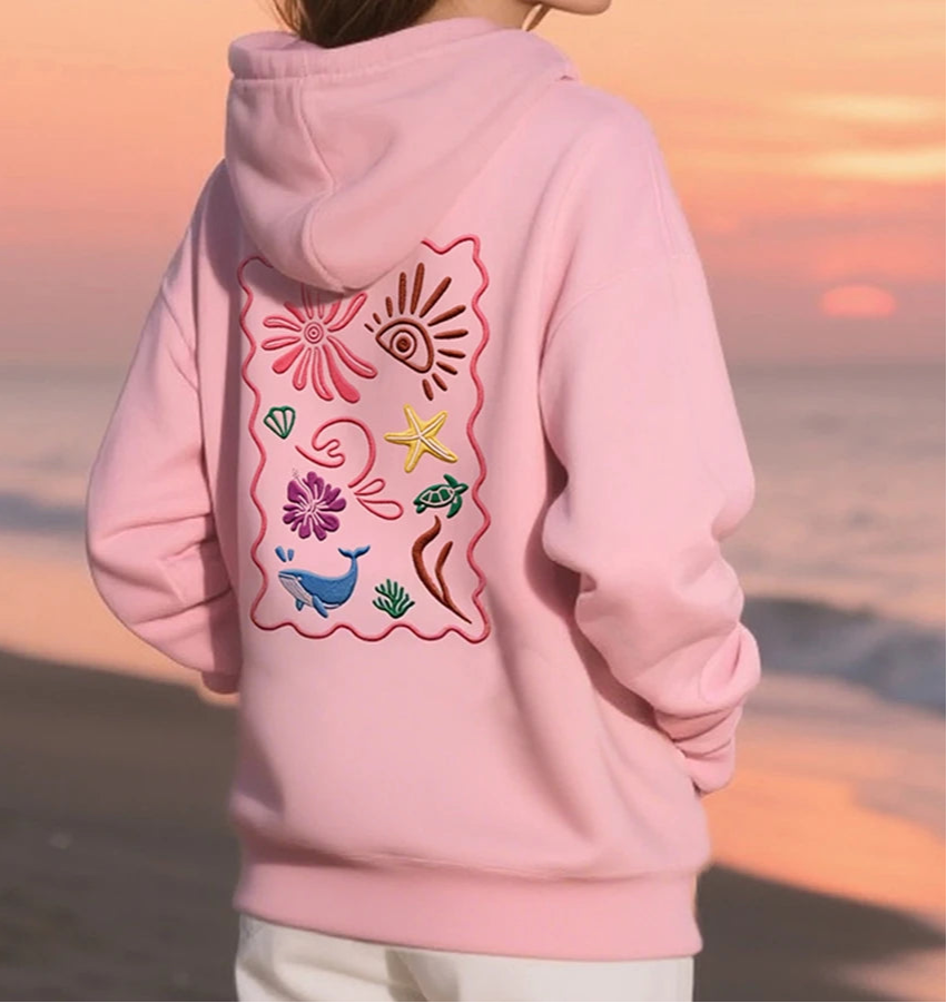 Person wearing a pink hoodie with embroidered designs on the back, standing on a beach at sunset.