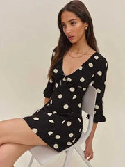 Woman wearing a black dress with white polka dots sitting on a chair against a plain background