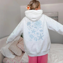 Person wearing a light gray hoodie with blue designs on the back, standing in a bedroom.