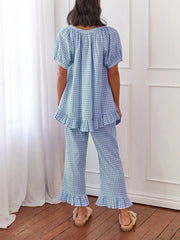 Person wearing a blue checkered outfit standing in a room with white curtains and wooden floor.