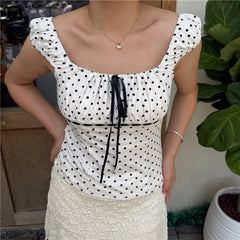 Woman wearing a white polka dot top with black dots, standing indoors next to a plant.