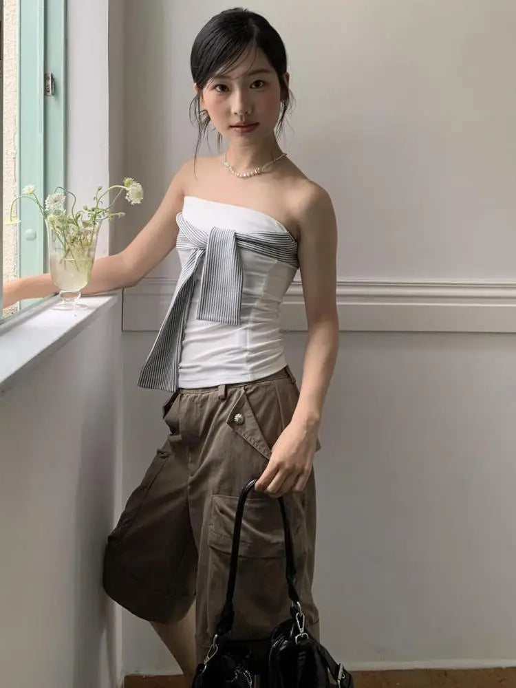Woman standing by a window holding a black bag, wearing a strapless top and brown pants.