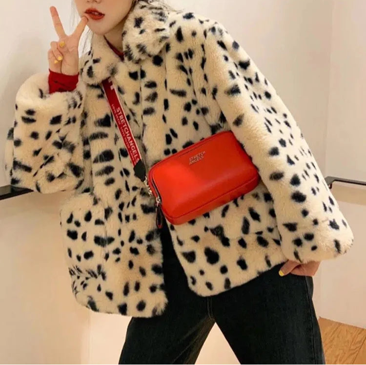 Person wearing a white coat with black spots, holding a red handbag, and making a peace sign.