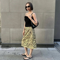 Woman wearing a black top and yellow snake print skirt standing against a gray wall.