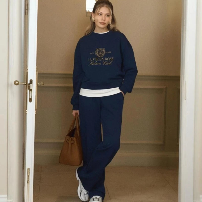 Woman wearing a navy blue sweatshirt and pants with a logo, standing in a room with beige walls.