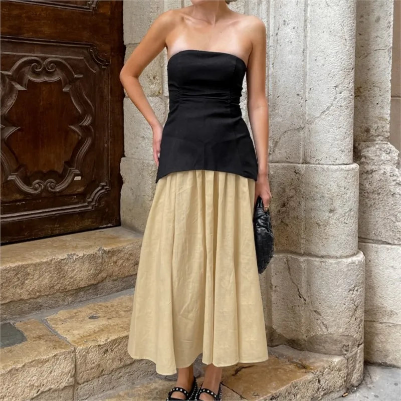 Woman wearing a black strapless top and beige skirt standing against a stone wall.