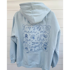 Light blue hoodie with a coastal-themed design on the back against a light wooden panel background