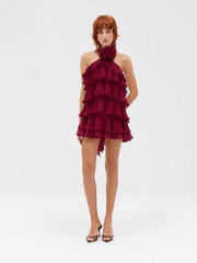 A model wearing a red halter-neck mini dress with dramatic tiered ruffles and a matching fabric flower collar, paired with brown strappy heels, against a plain white studio background.