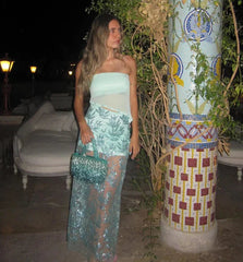 Woman in a light blue strapless top and green lace skirt standing next to a decorative column at night.