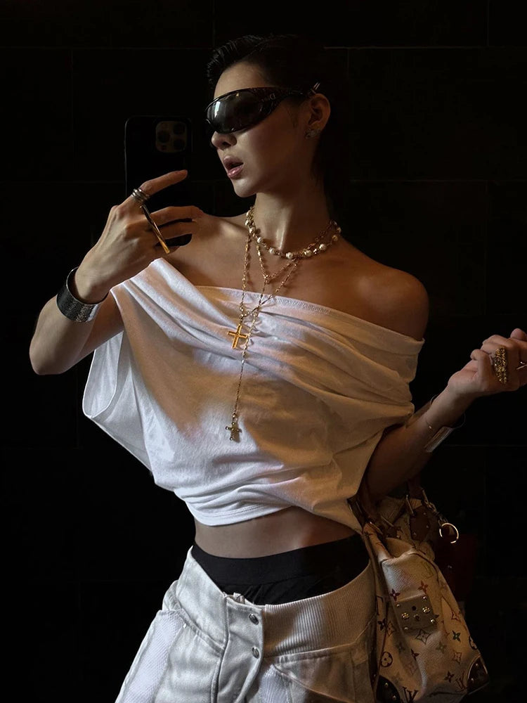 Person wearing a white off-shoulder top and sunglasses, holding a phone and a bag.