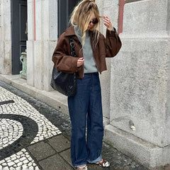Woman in brown jacket and blue jeans standing against a stone wall.
