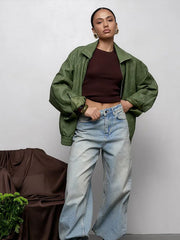 Person wearing a green jacket, brown top, and blue jeans against a plain background