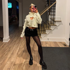 Person taking a mirror selfie wearing a white cropped sweater with heart designs, black shorts, and black shoes in a home setting.
