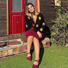 Woman sitting on a wooden bench in front of a red door with floral decorations.