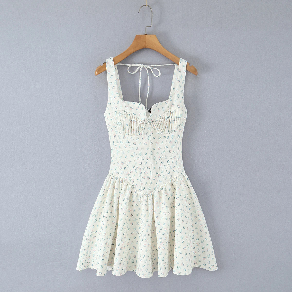 A sleeveless white mini dress with a small green and blue floral print, hanging on a wooden hanger against a solid gray background.