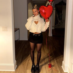 Person wearing a white sweater with a heart cutout, holding red heart balloons in a room.