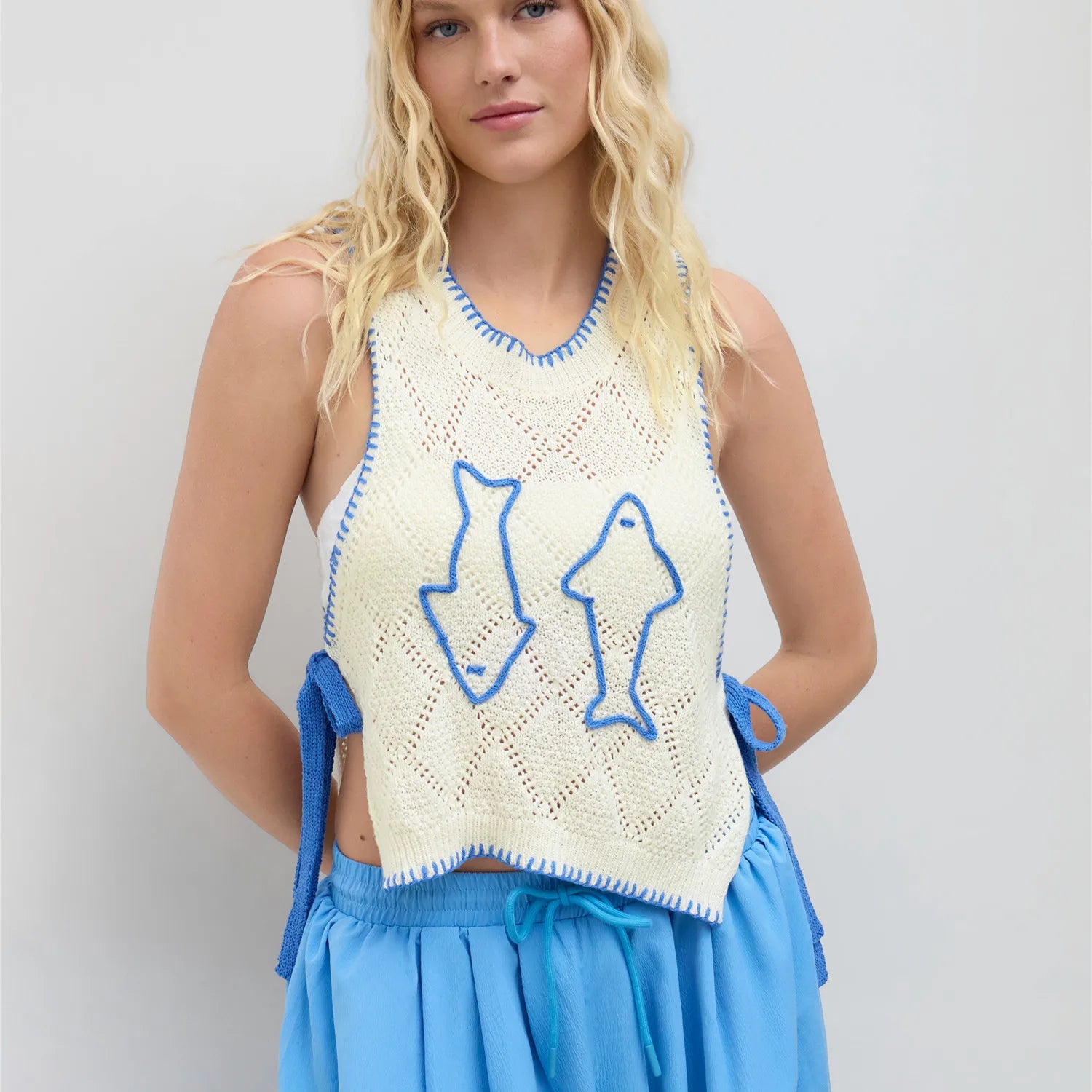 Woman wearing a crochet top with fish designs and blue shorts on a light gray background