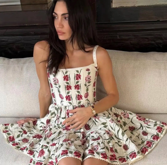 A woman sits on a cream-colored sofa wearing a sleeveless cream dress with a red and green floral print.